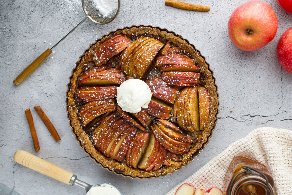 Overhead view of a baked apple tart in a round pan, topped with a scoop of vanilla ice cream and powdered sugar. Surrounding the tart are whole apples, cinnamon sticks, a jar of honey, and a sifter with powdered sugar.