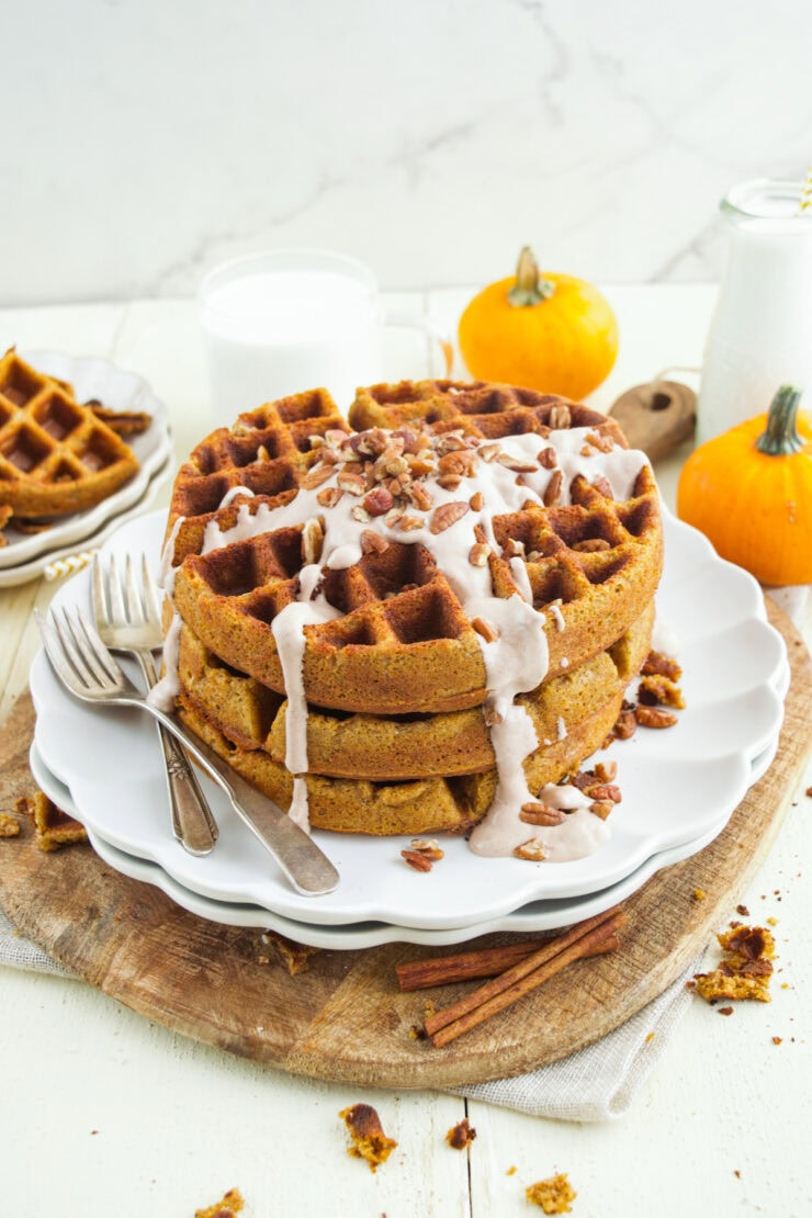 A stack of butternut squash waffles drizzled with icing and topped with pecans sits on a white plate with forks. Small pumpkins and glasses of milk are in the background on a wooden board.