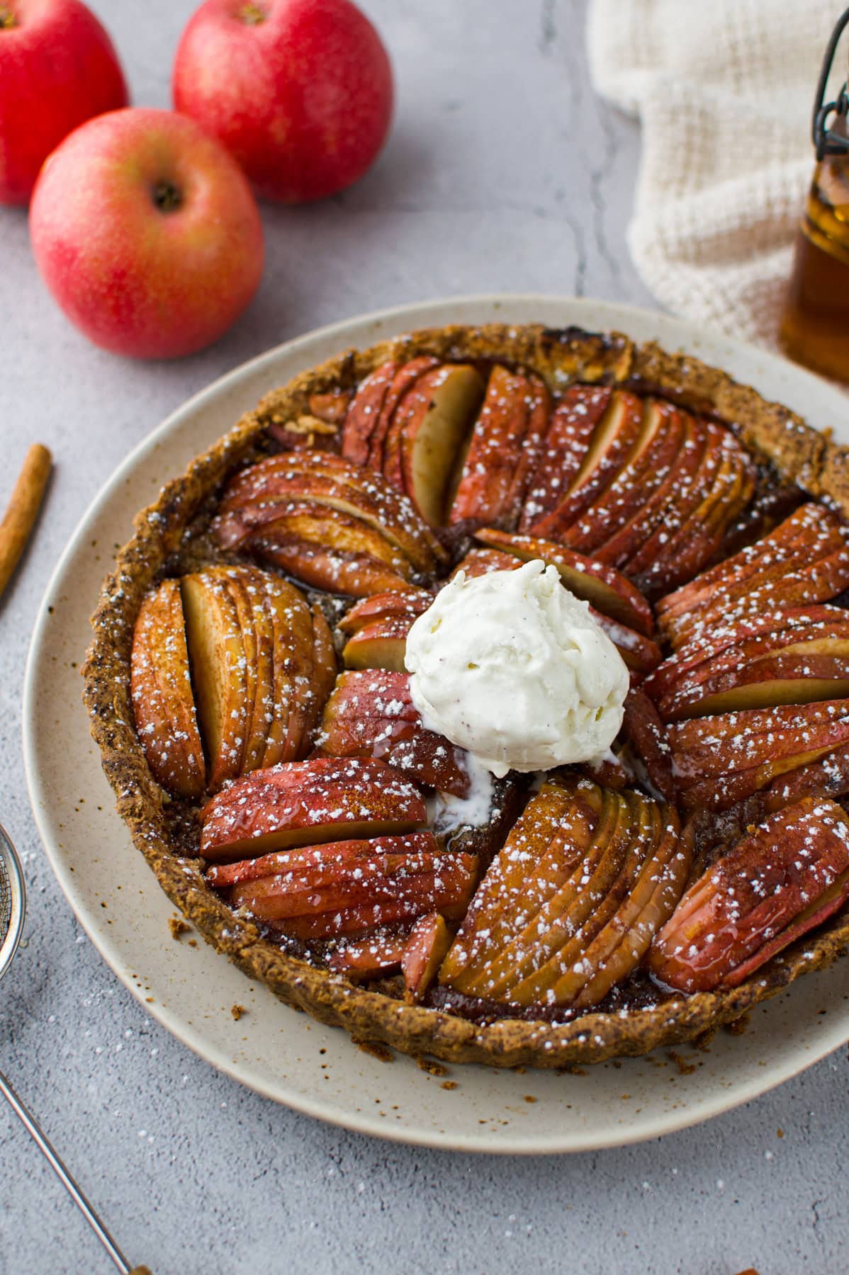 A baked apple tart with sliced apples arranged in a circular pattern, topped with a scoop of vanilla ice cream and powdered sugar, sits on a plate. Three red apples and a small jar are in the background.