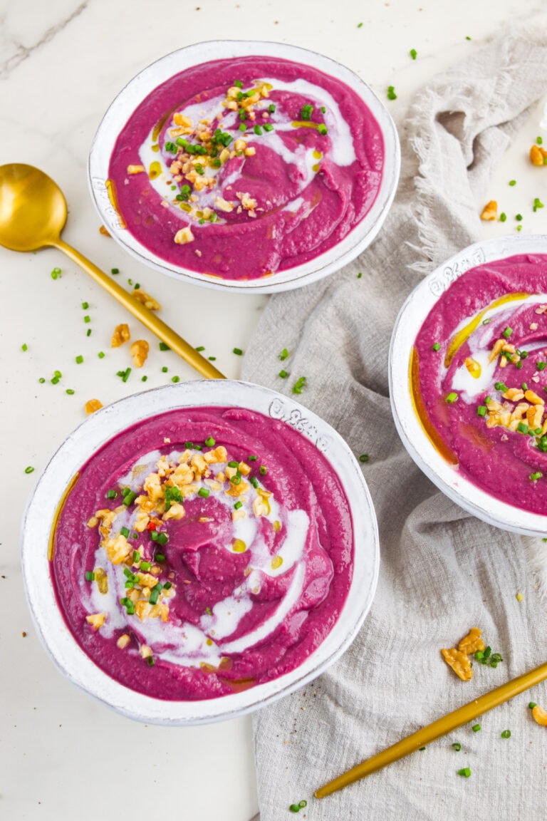 Close-up of a vibrant magenta beet soup served in a white bowl, swirled with coconut cream and topped with chopped walnuts and fresh chives, with a gold spoon and linen napkin on the side.
