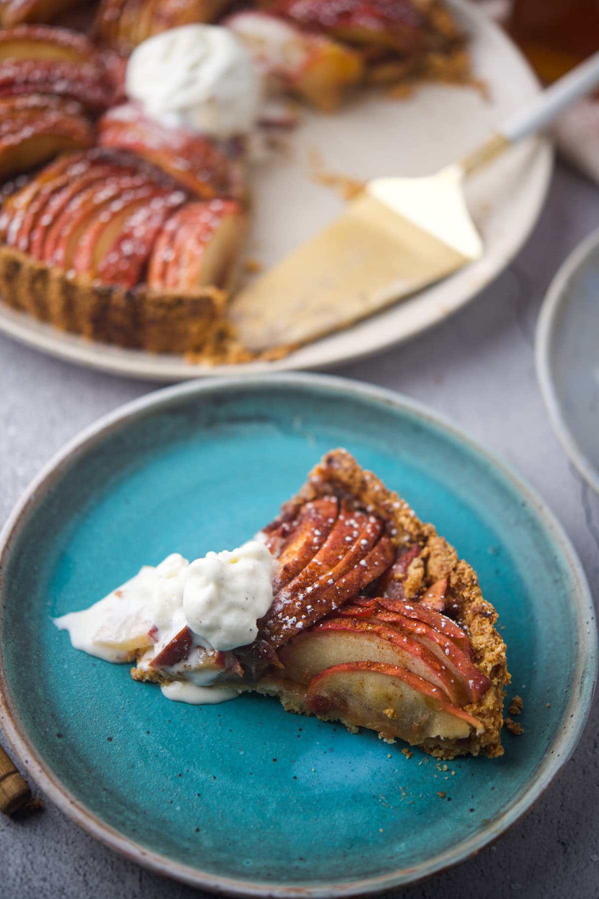 A slice of apple tart with a crumbly crust and a scoop of vanilla ice cream is served on a blue plate, with the rest of the tart and a pie server visible in the background.