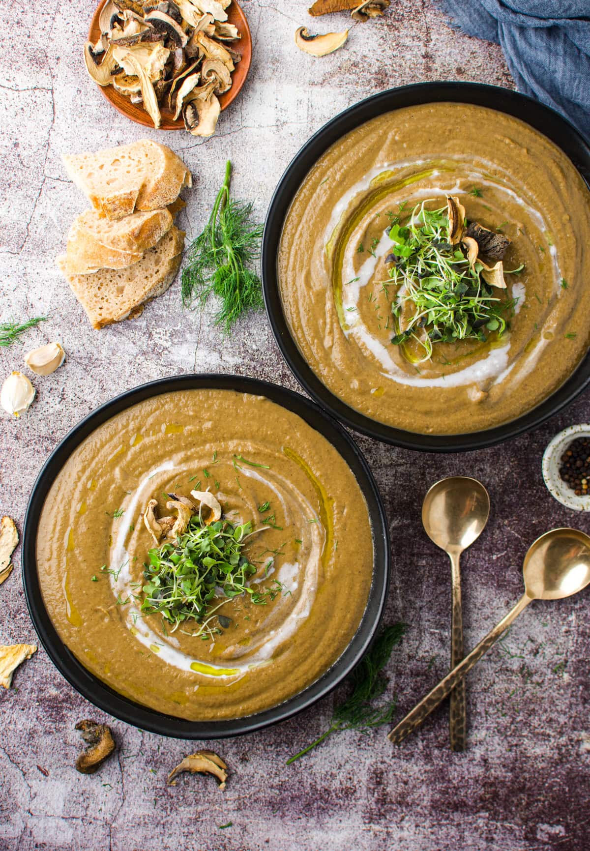 Two bowls of creamy mushroom soup topped with microgreens and a drizzle of oil, surrounded by sliced bread, dried mushrooms, fresh dill, garlic, and two gold spoons on a rustic surface.