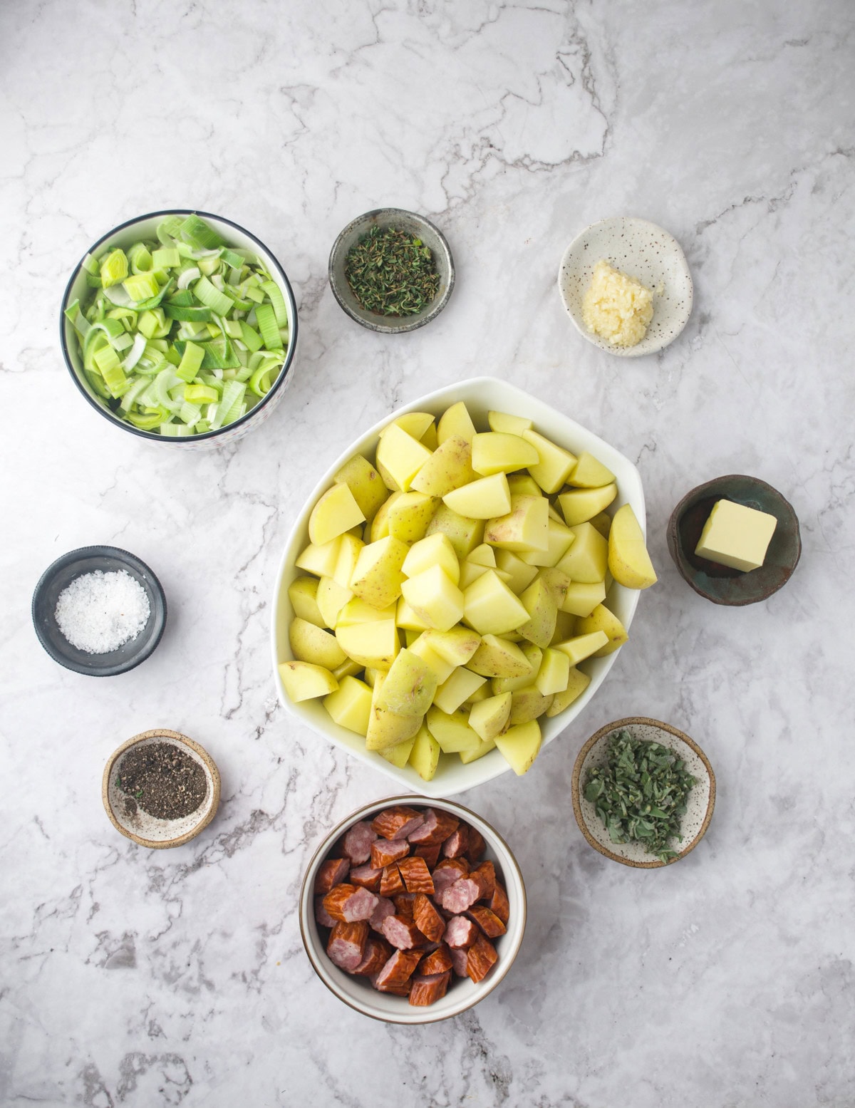 An overhead view of small bowls containing chopped potatoes, sliced leeks, sausage pieces, minced garlic, fresh herbs, butter, salt, and pepper, all arranged on a marble countertop.