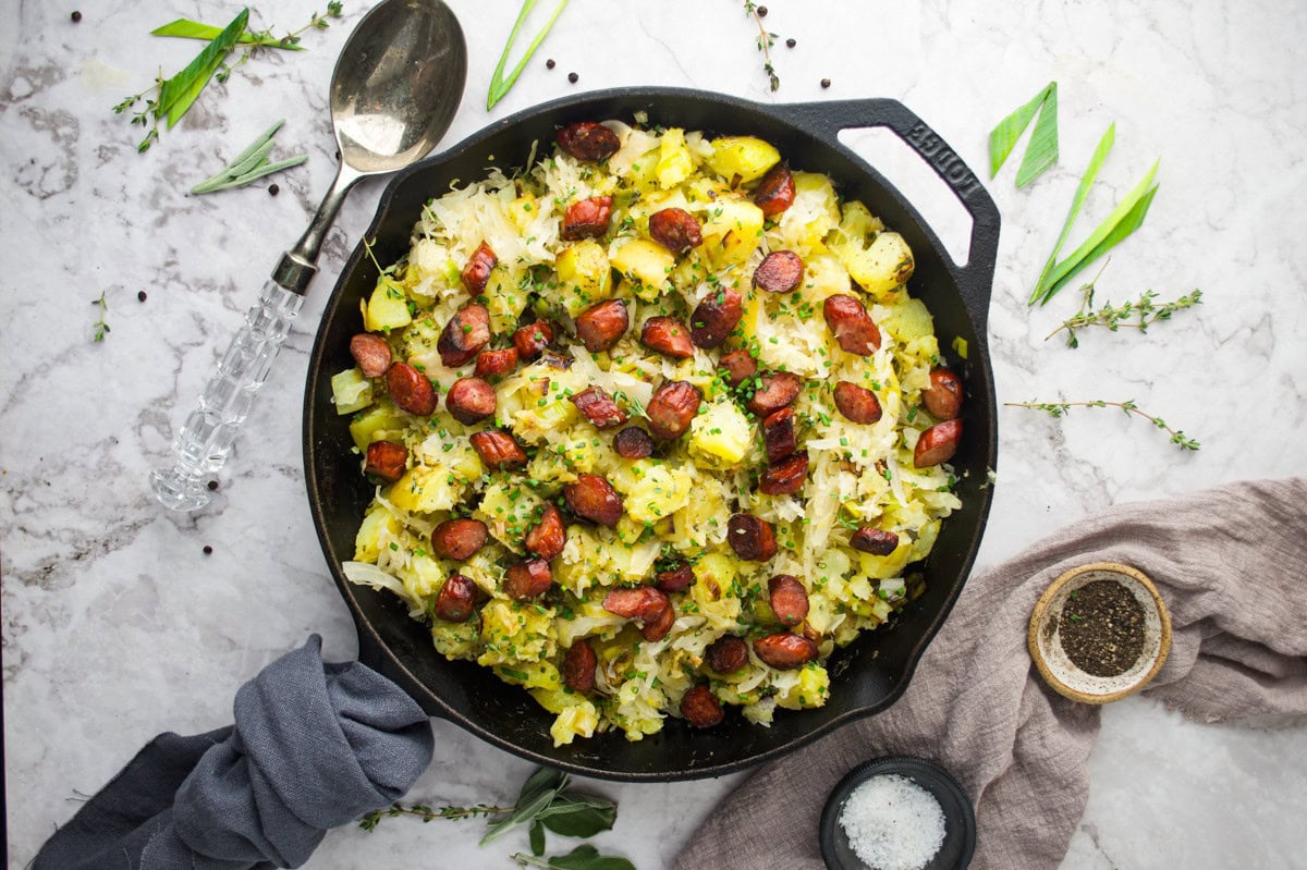 A cast iron skillet filled with roasted potatoes, sliced sausage, sauerkraut, and herbs sits on a marble surface, surrounded by a spoon, salt, pepper, and sprigs of fresh herbs.