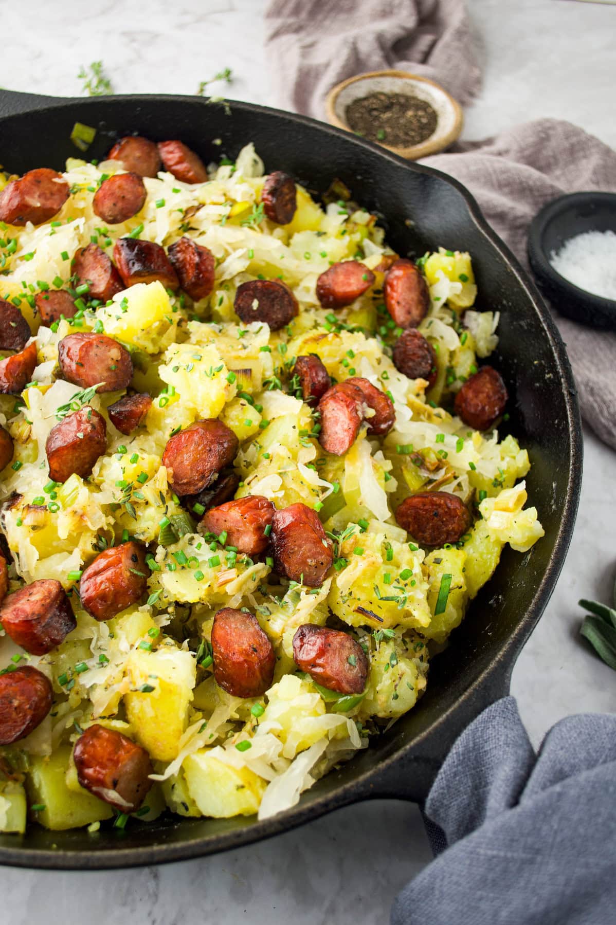 A skillet filled with saut&eacute;ed potatoes, sliced sausage, and sauerkraut, garnished with chopped chives, sits on a marble surface with salt, pepper, and a gray napkin nearby.