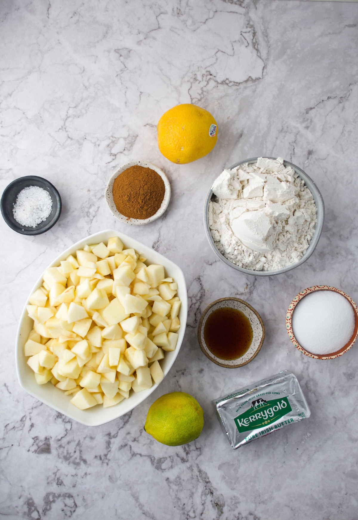 A marble countertop displays bowls of chopped apples, flour, sugar, cinnamon, flaky salt, a stick of butter, a lemon, a lime, and a small bowl of vanilla extract.