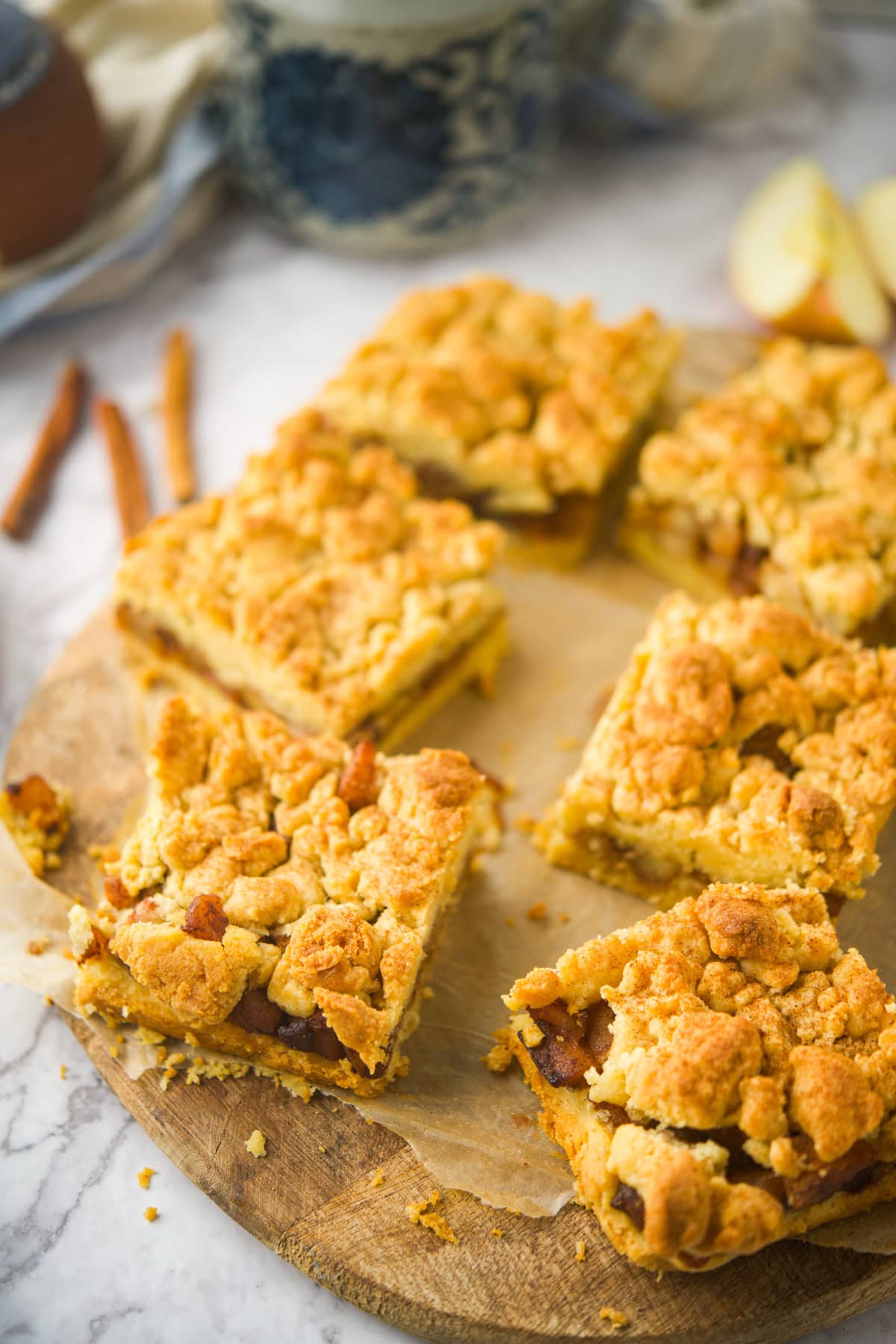 Six golden, crumbly apple bars are arranged on a round wooden board lined with parchment paper. Cinnamon sticks and apple slices are visible in the background. The bars have a textured, rustic topping.