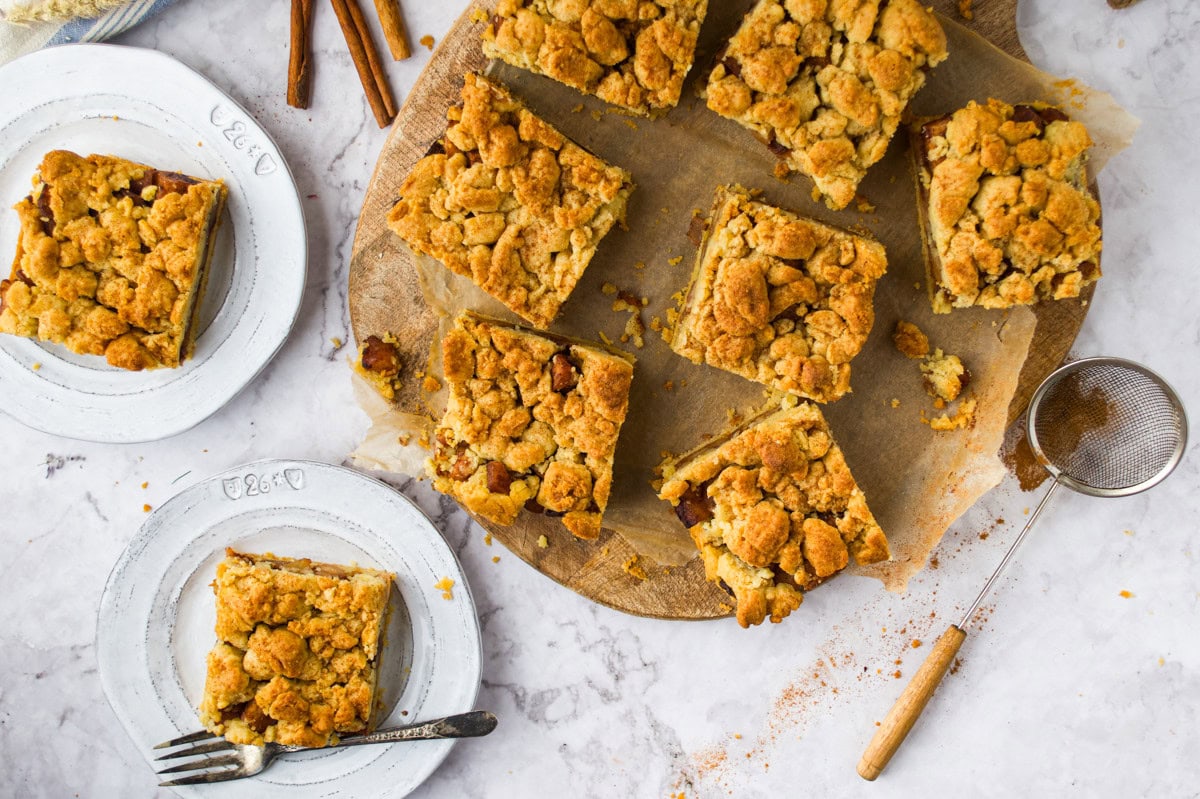 Top-down view of crumbly apple pie bars on parchment paper, set on a round wooden board, with two servings plated nearby. A fork, cinnamon sticks, and a fine mesh strainer with cinnamon are also visible.