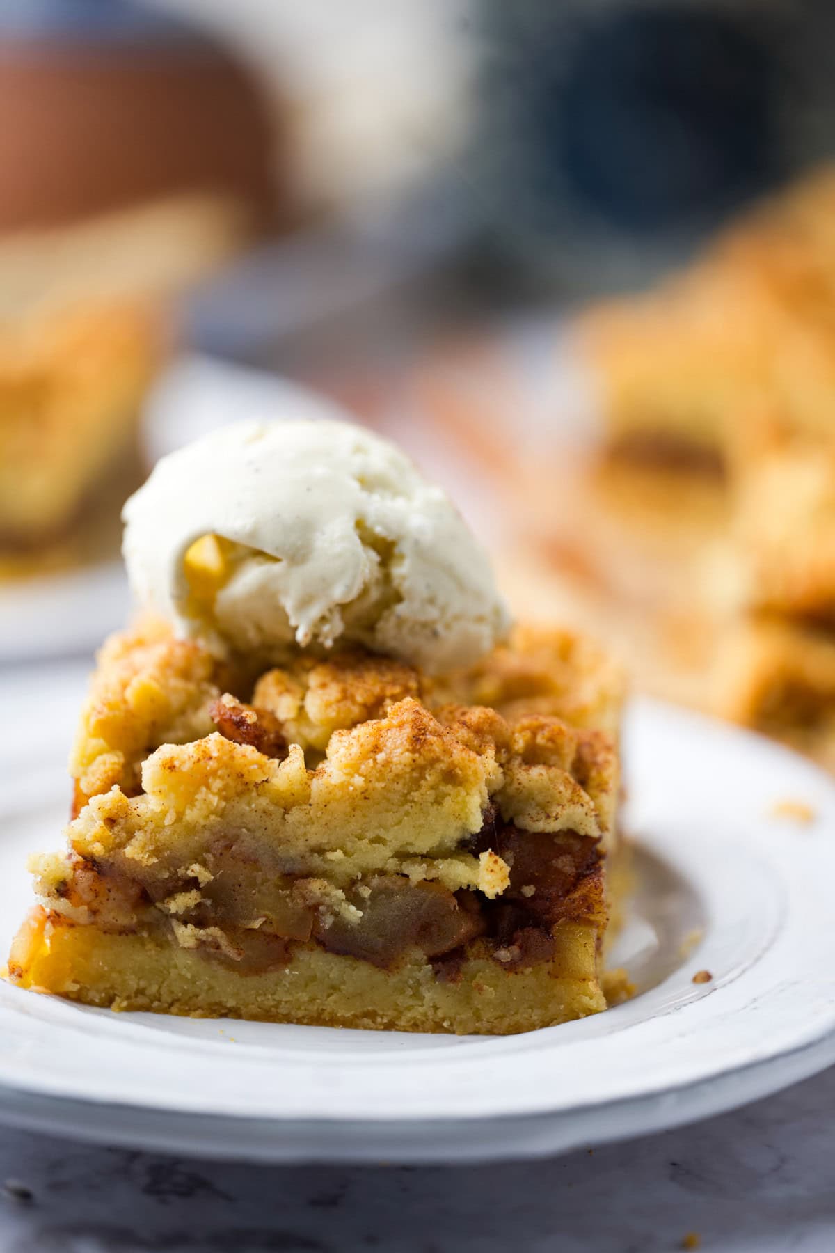 A close-up of a slice of apple crumb pie bars topped with a scoop of vanilla ice cream, served on a white plate. The pie has a golden, crumbly topping and visible apple filling.