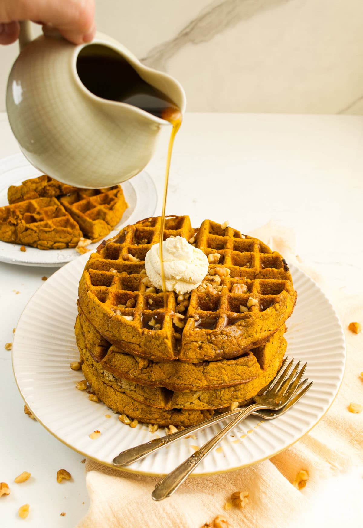A hand pours syrup from a small pitcher onto a stack of Tiger Nuts And Pumpkin Waffles topped with whipped cream and chopped nuts on a white plate, with two forks nearby and another plate of waffles in the background.