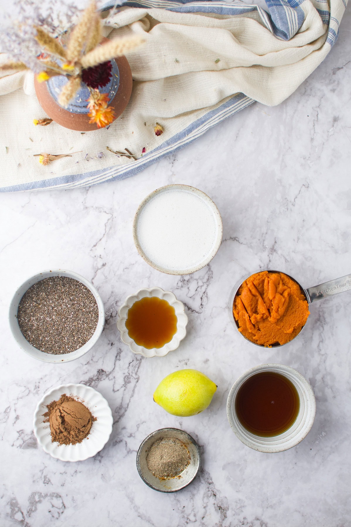 A flat lay of various baking ingredients on a marble surface, including chia seeds, sugar, pumpkin puree, vanilla extract, cocoa powder, a lemon, black pepper, and a cup of liquid, with a vase of dried flowers above.
