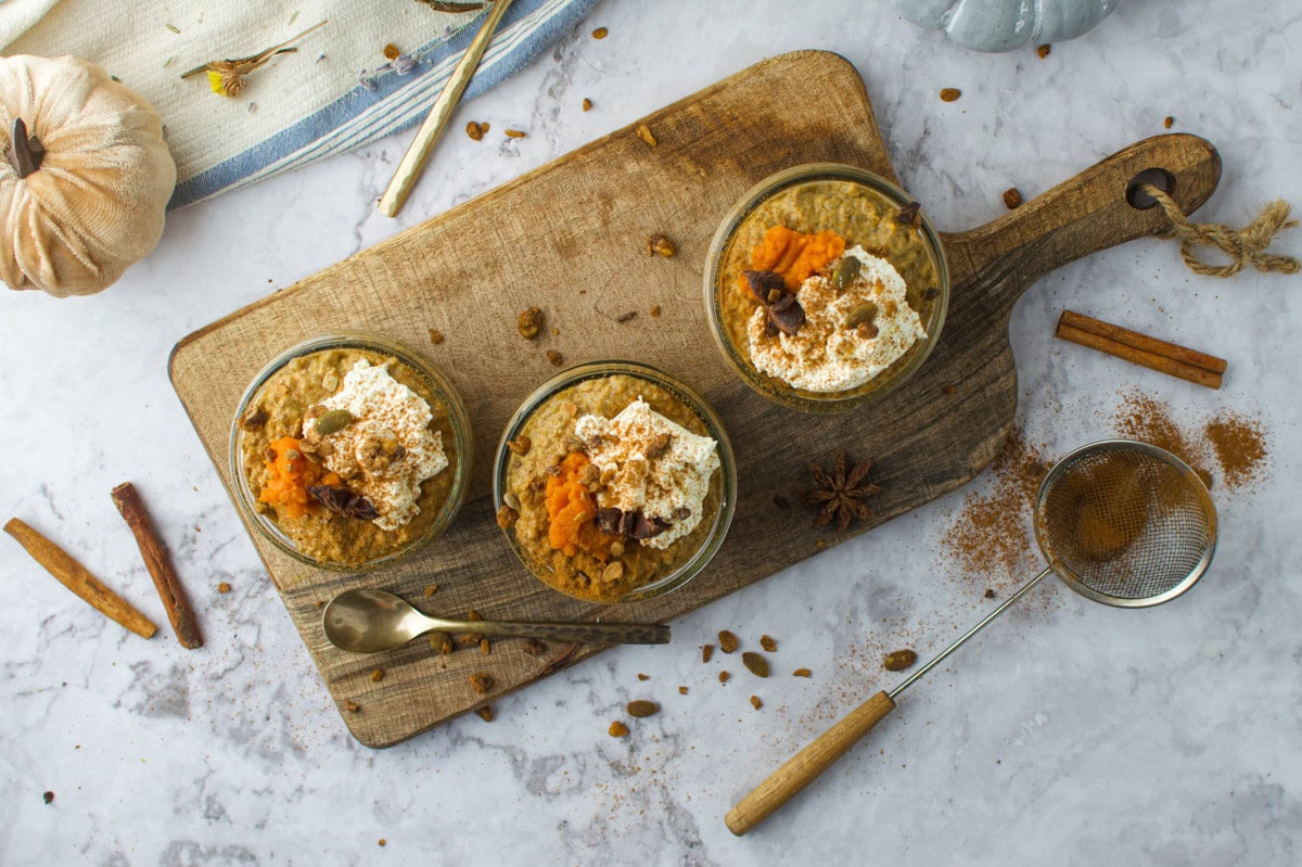 Three glass jars filled with dessert, topped with whipped cream and pumpkin, sit on a wooden board. Cinnamon sticks, a spice sieve, and star anise are scattered on a marble surface nearby.