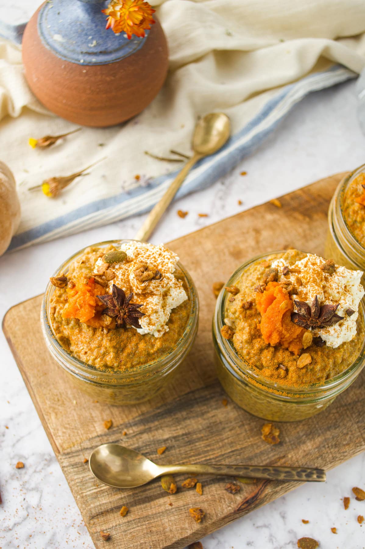 Two small jars of pumpkin chia pudding topped with whipped cream, granola, star anise, and pumpkin puree sit on a wooden board. A gold spoon, cloth, and ceramic pot are in the background.