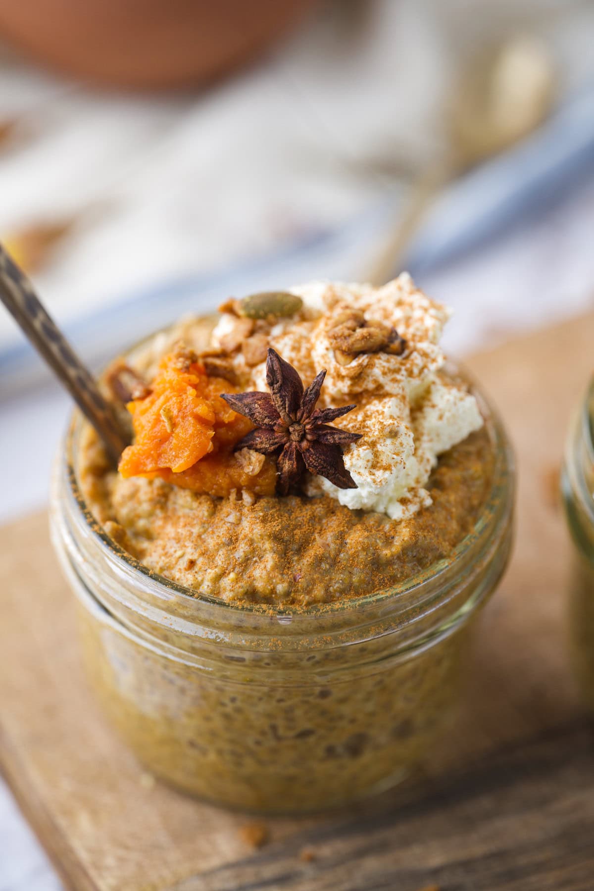 A glass jar filled with pumpkin chia seed pudding dessert topped with whipped cream, granola, a spoonful of pumpkin puree, a star anise, and a sprinkle of spice, sitting on a wooden surface.