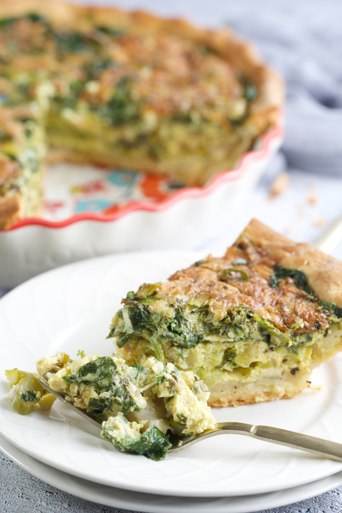 A close-up of a slice of Gluten-Free Asparagus Leek And Spinach Quiche on a white plate with a fork, in front of a larger quiche in a pie dish. The quiche filling appears to include spinach and cheese.