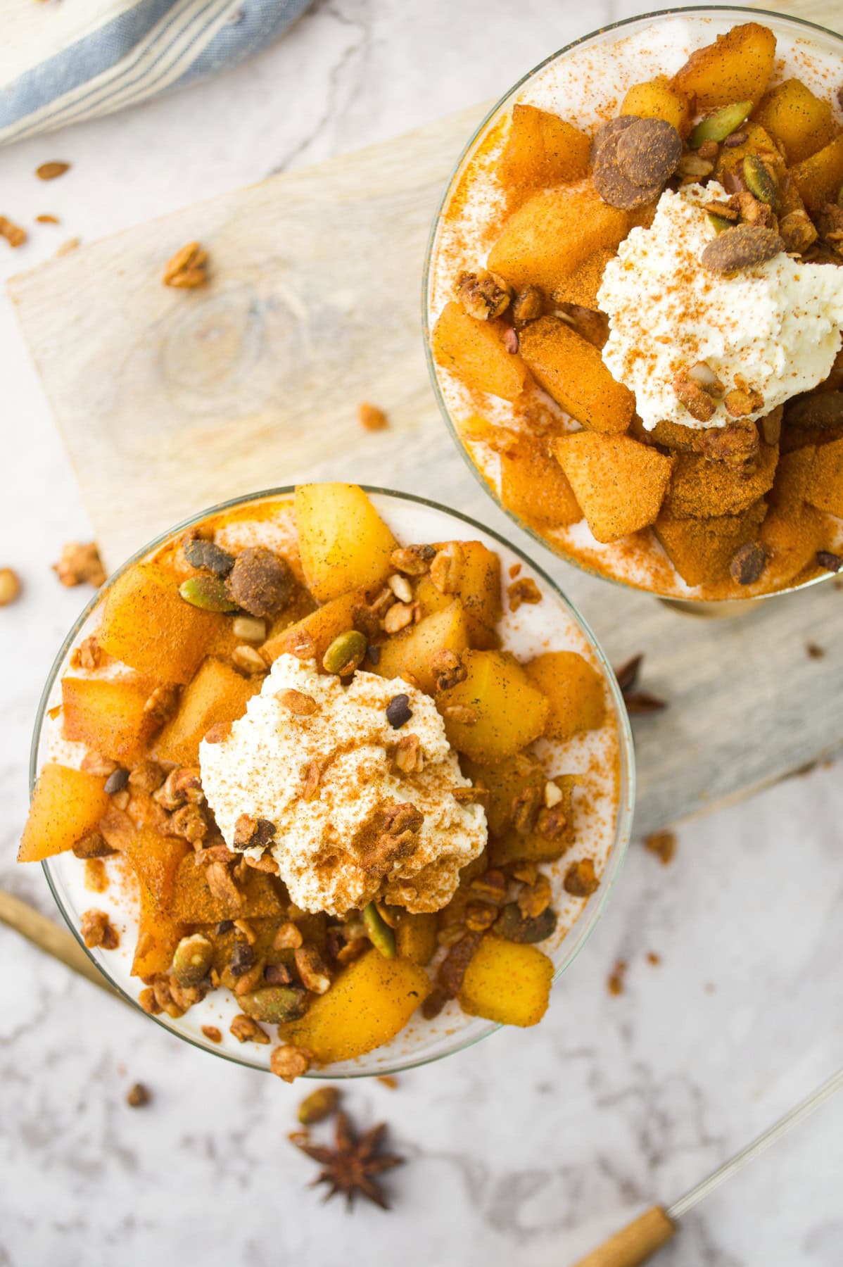 Two glass bowls filled with spiced baked apple slices, whipped cream, granola, and a sprinkling of cinnamon, placed on a light wooden board with star anise and granola scattered nearby.