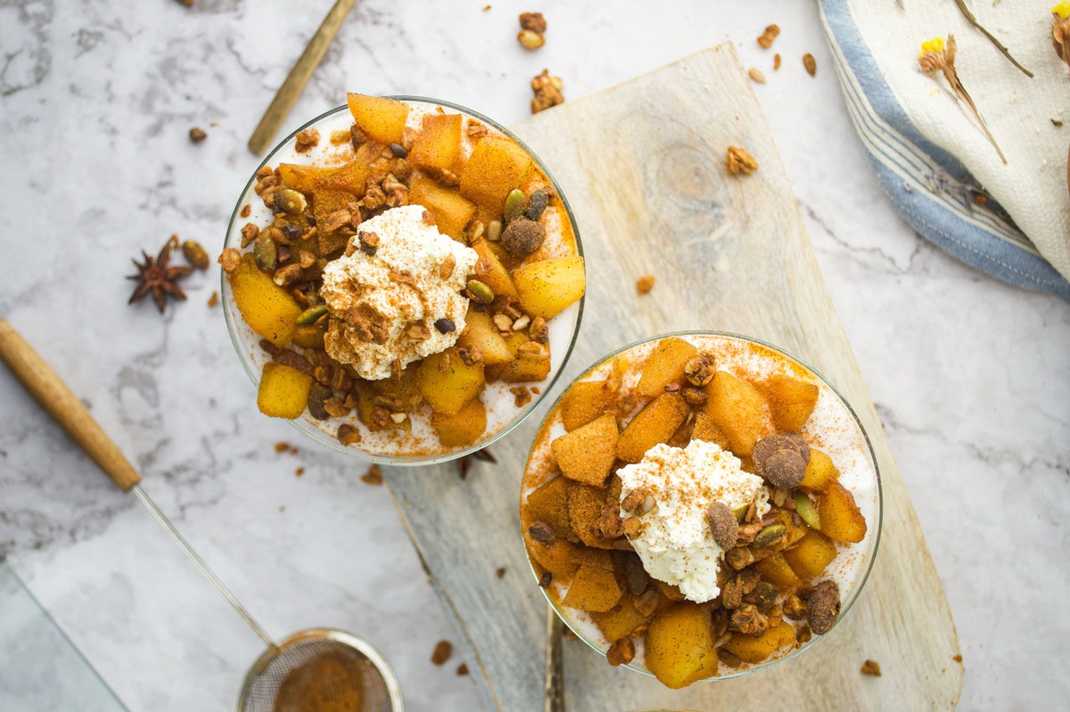 Two glass bowls filled with spiced apple chunks, granola, and whipped cream sit on a wooden board. A sieve with cinnamon and a napkin with dried flowers are nearby on a light marble surface.