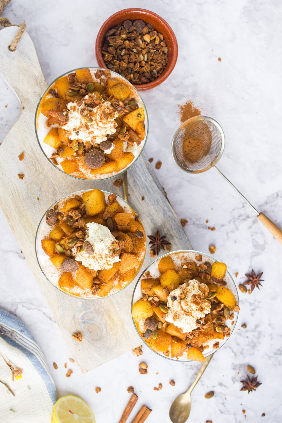 Three bowls of dessert topped with whipped cream, spiced apples, granola, and cinnamon sit on a wooden board. A small bowl of granola, a sieve with cinnamon, and star anise are scattered on a marble surface.
