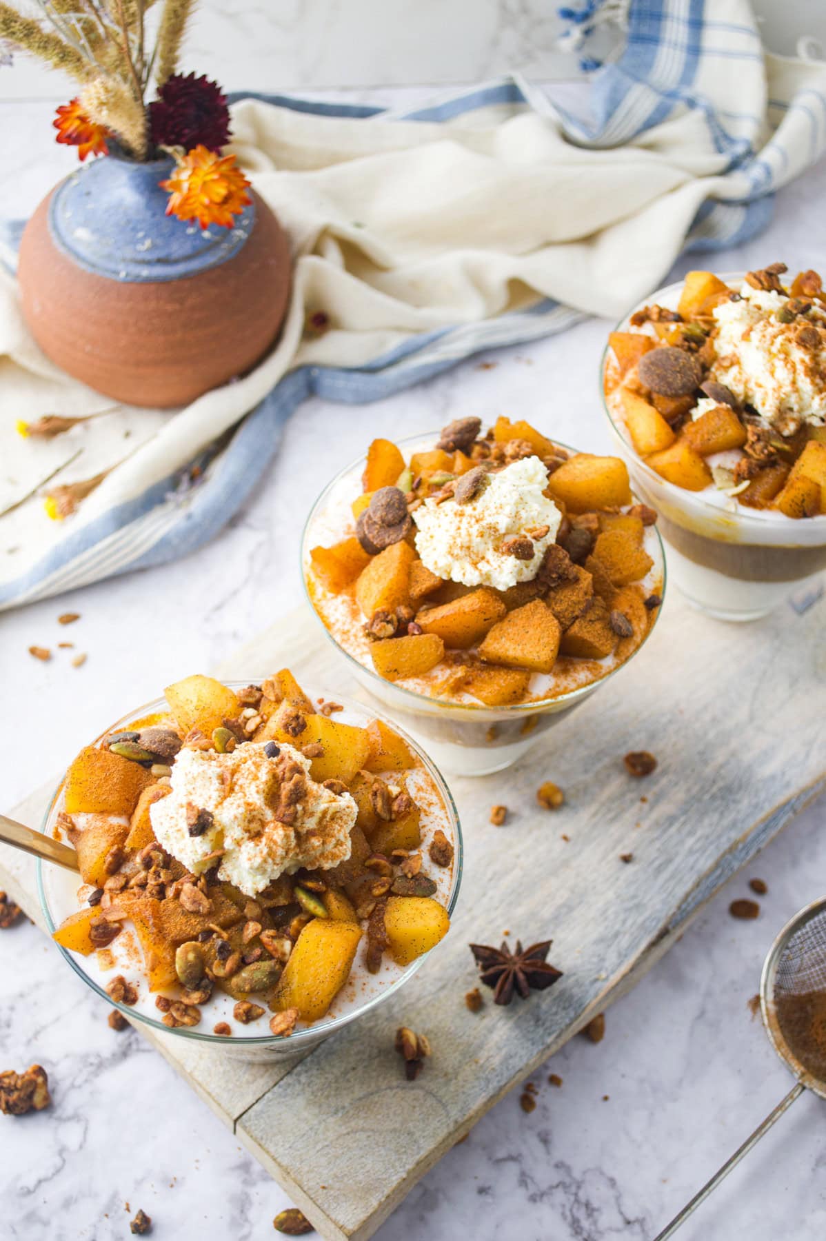Three glass bowls filled with yogurt, caramelized apple chunks, granola, and whipped cream sit on a wooden board. Dried flowers in a vase and a cloth napkin decorate the background.