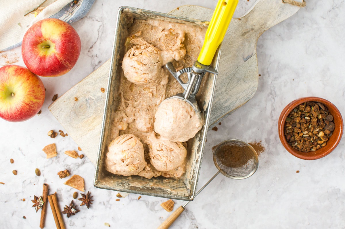 A metal loaf pan holds several scoops of cinnamon ice cream with a yellow-handled scooper. Nearby are two apples, granola, cinnamon sticks, and waffle pieces on a marble surface.