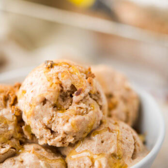 Three scoops of creamy, apple cinnamon-spiced ice cream with a drizzle of honey sit in a white bowl, with a textured, nutty topping visible. The background shows a blurred ice cream container and scoop.