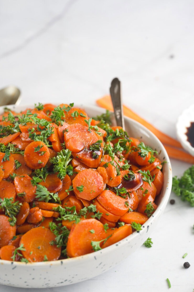 A bowl filled with sliced cooked carrots garnished with fresh chopped parsley. Spoons are placed in the bowl, and a carrot and herbs are visible in the background on a white surface.