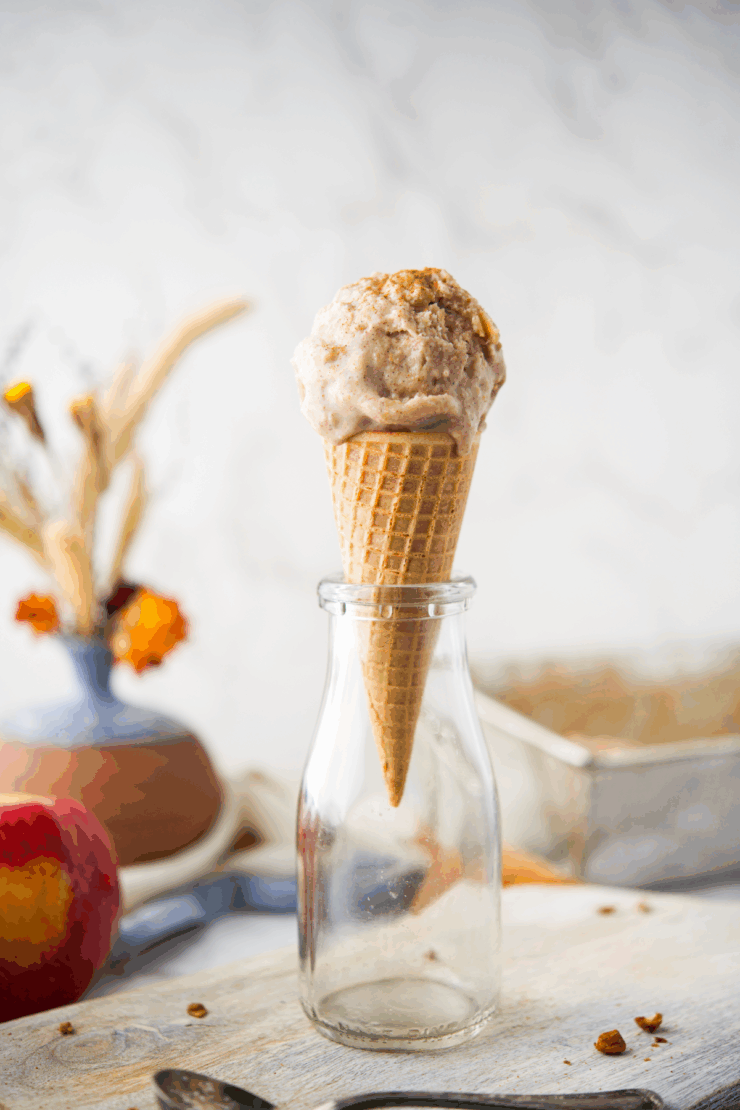 A scoop of apple cinnamon ice cream in a waffle cone is placed upside down in a glass milk bottle. In the blurred background, there’s a vase with dried flowers, an apple, and a metal tray.