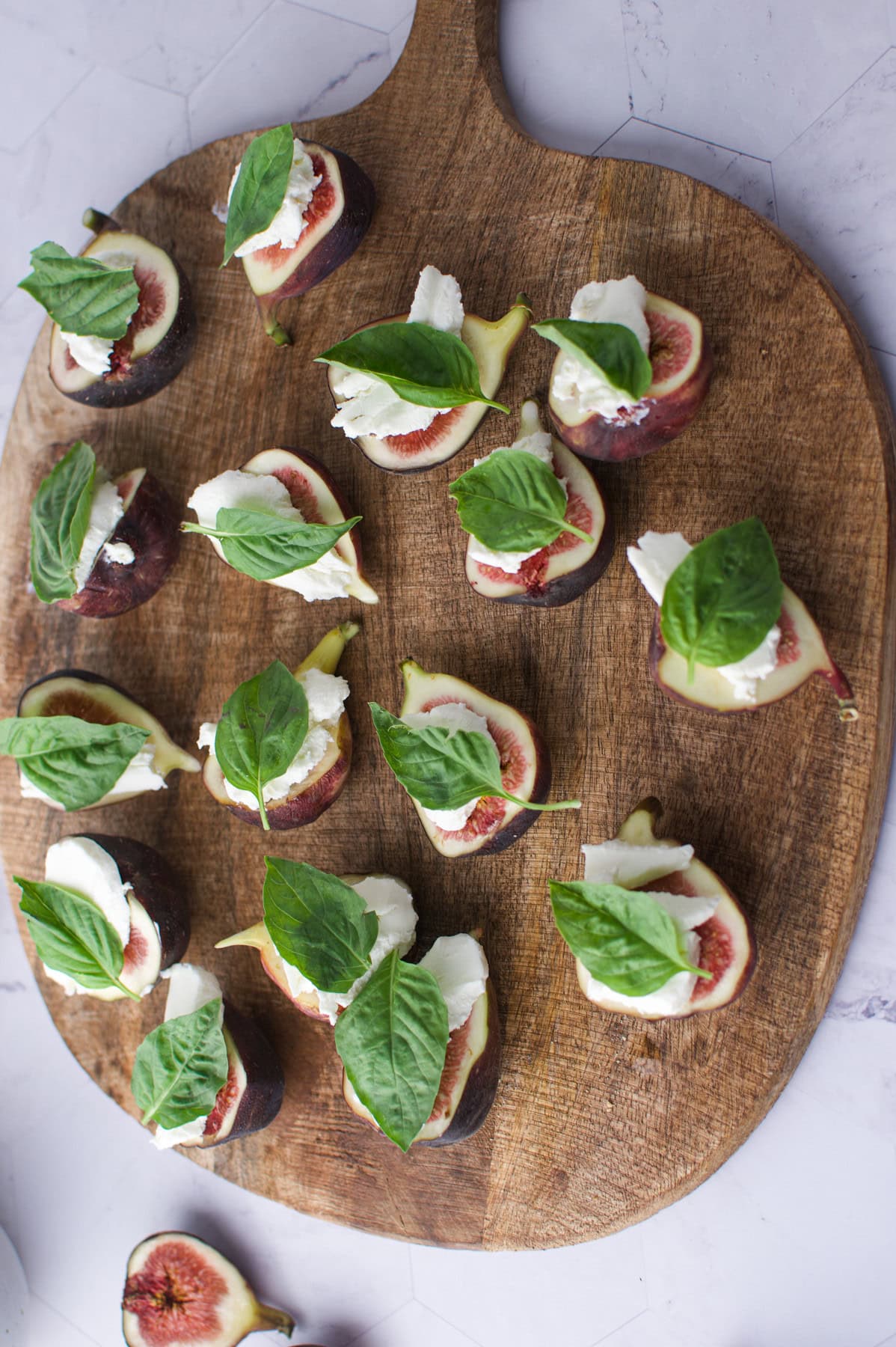 Sliced figs topped with mozzarella cheese and fresh basil leaves are arranged on a wooden serving board, set against a light marble background.