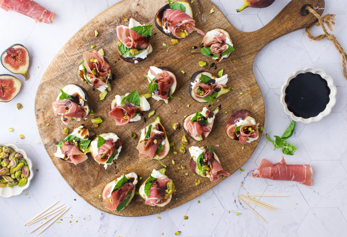 A wooden serving board topped with small appetizers made of prosciutto, mozzarella, basil, and figs. Pistachios are scattered around, with a bowl of balsamic vinegar and more ingredients on a marble surface.