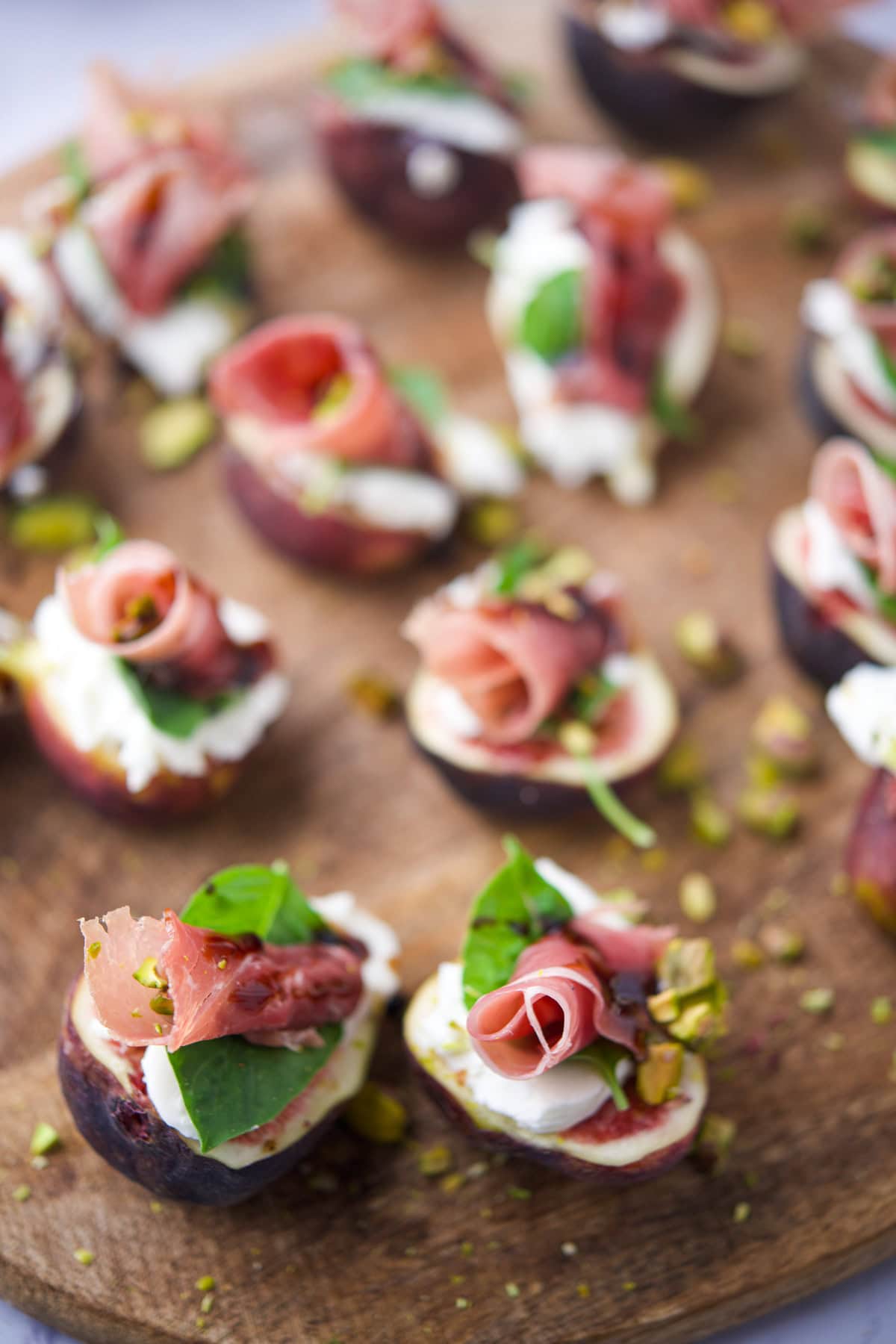 Close-up of appetizers on a wooden board, featuring halved figs topped with creamy cheese, prosciutto, fresh basil leaves, and chopped pistachios. The snacks are arranged in a scattered, inviting manner.