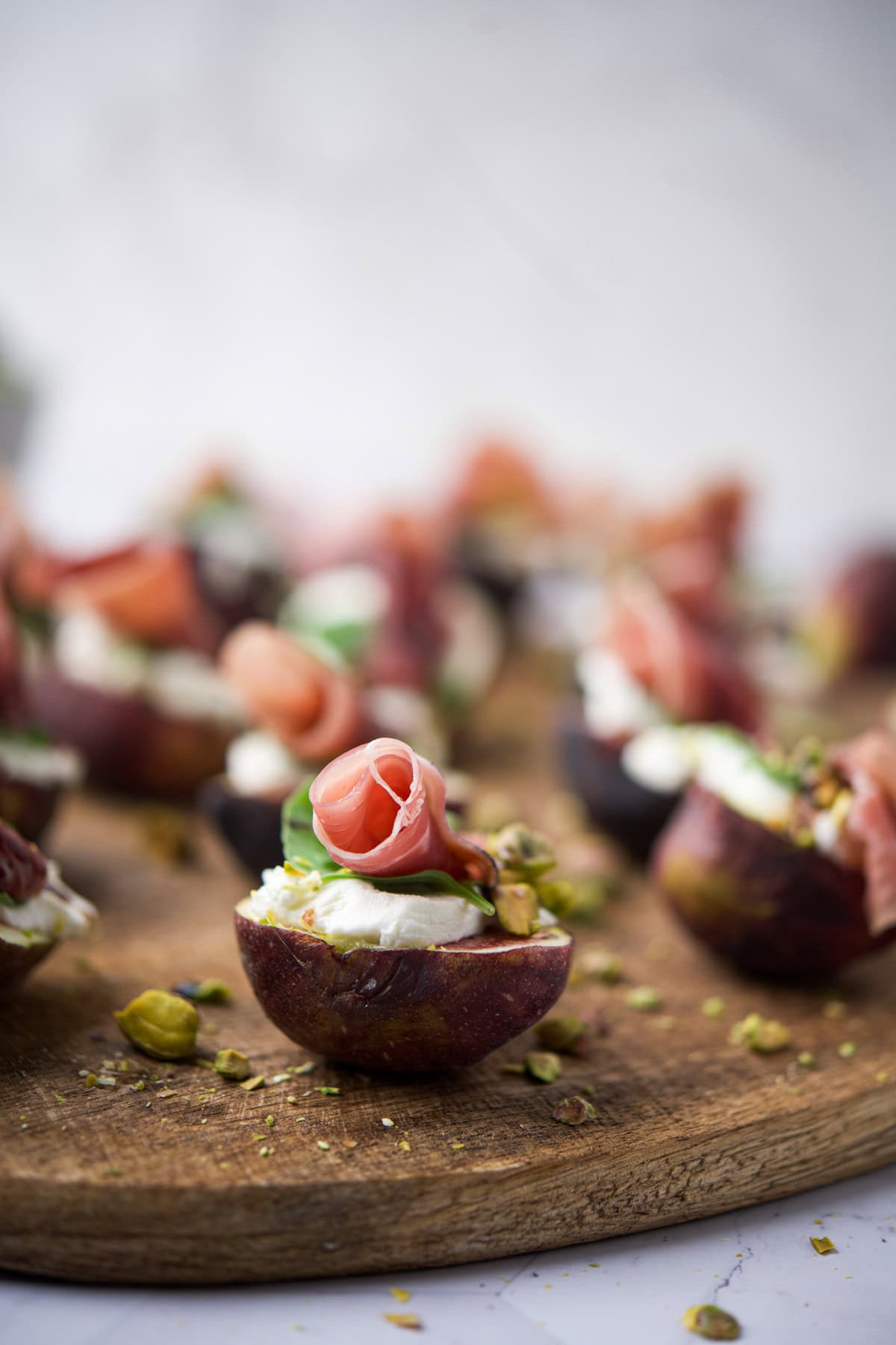 Halved figs topped with creamy cheese, rolled prosciutto, and chopped pistachios are arranged on a wooden serving board, with a blurred background of more appetizers.