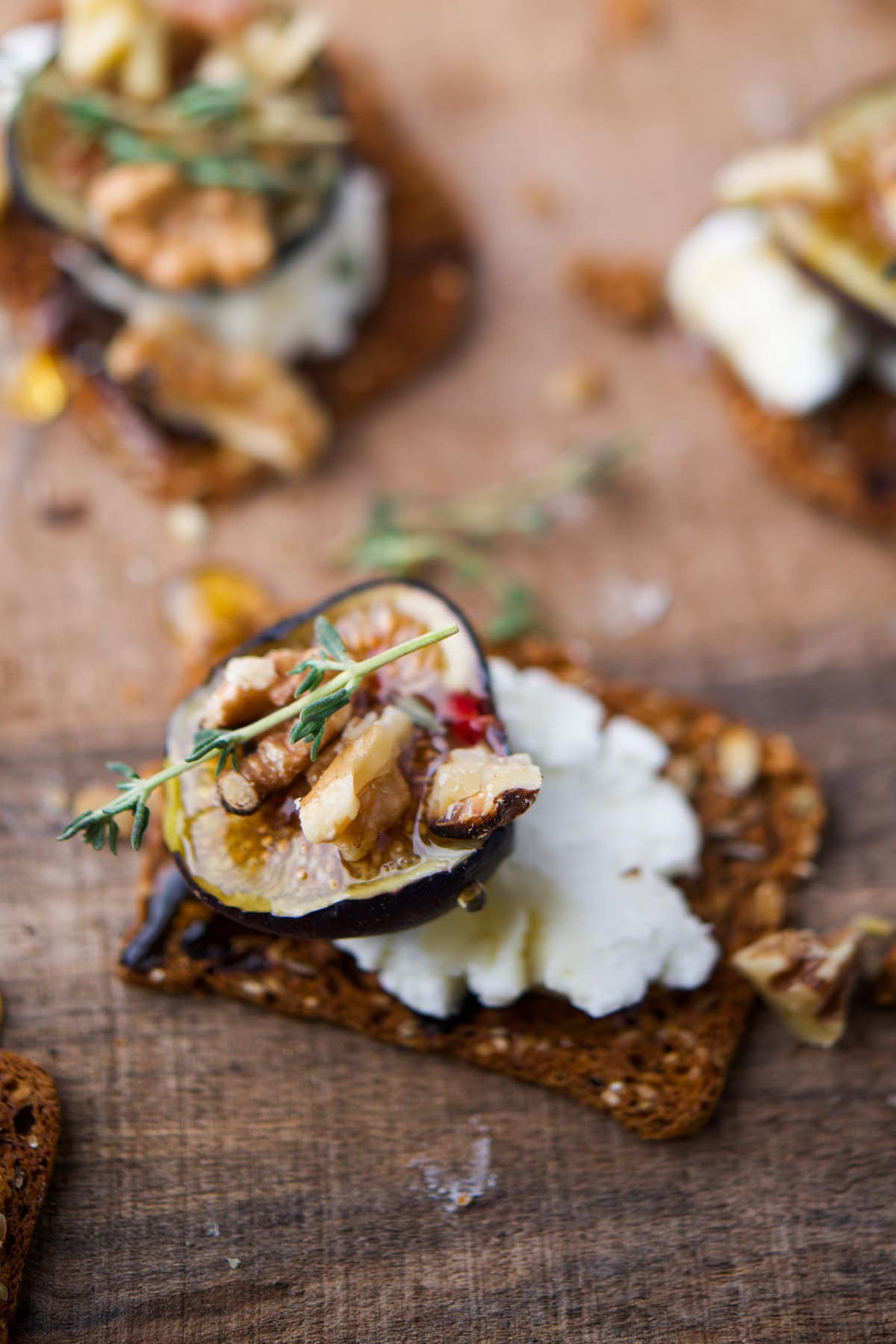 A close-up of a cracker topped with goat cheese, sliced fig, walnut pieces, fresh thyme, and a drizzle of honey on a wooden surface.