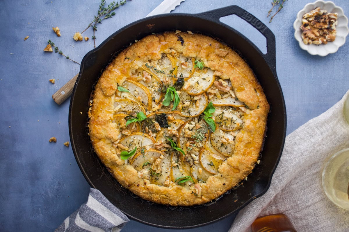 A rustic savory tart with sliced pears, herbs, and walnuts sits in a black cast-iron skillet on a blue surface, surrounded by a knife, a small plate of walnuts, and a linen napkin.