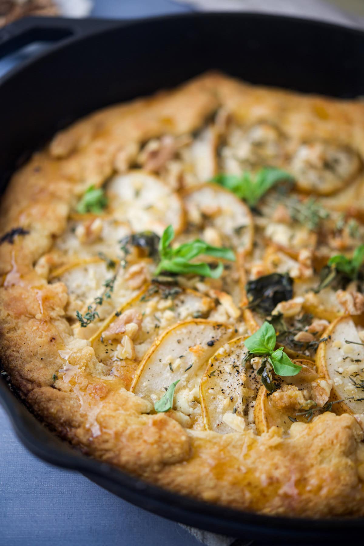 A close-up of a rustic pear and cheese tart garnished with fresh herbs, baked in a black cast iron skillet. The golden crust surrounds thinly sliced pears and crumbled cheese.