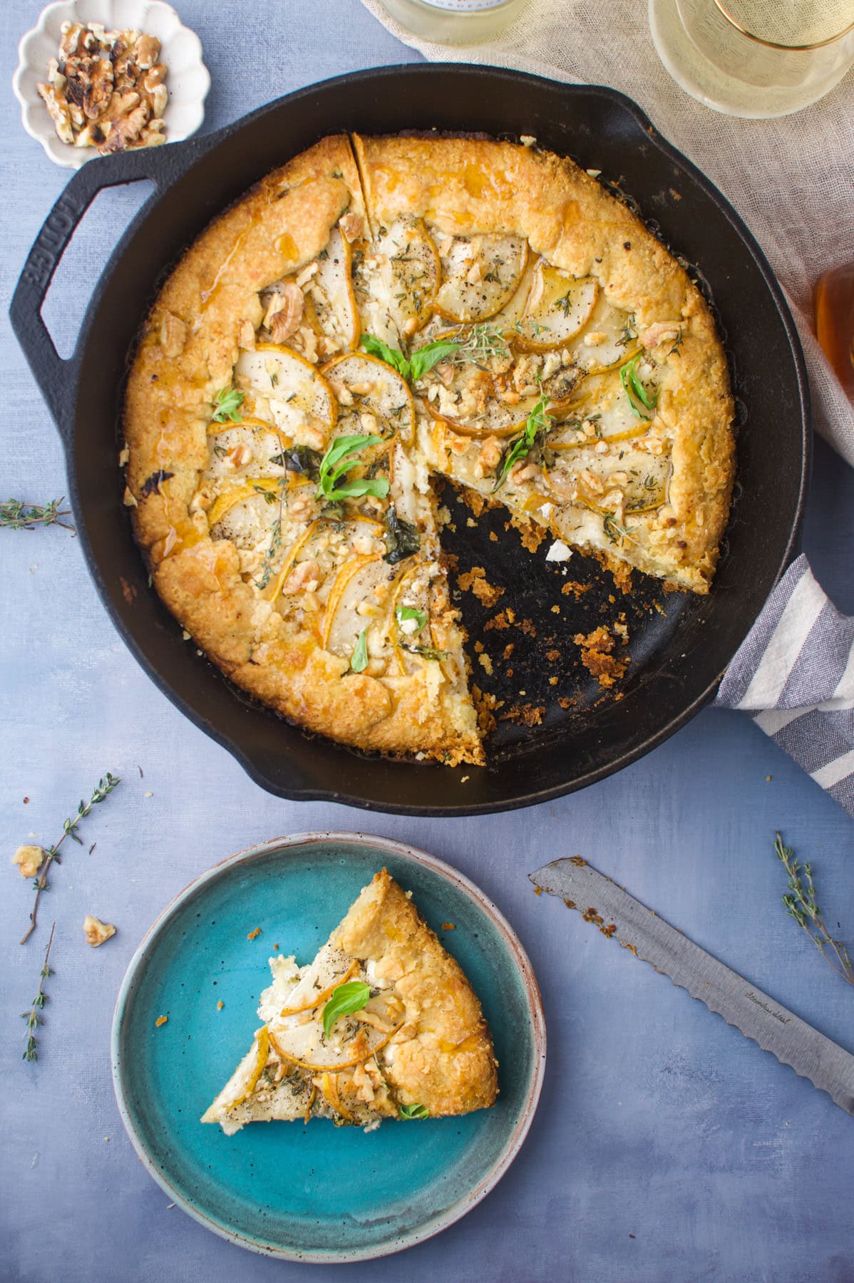 A golden, rustic savory pie with herbs, baked in a cast iron skillet, sits on a blue surface. One slice is served on a blue plate nearby, and a serrated knife and a small dish of walnuts are visible.