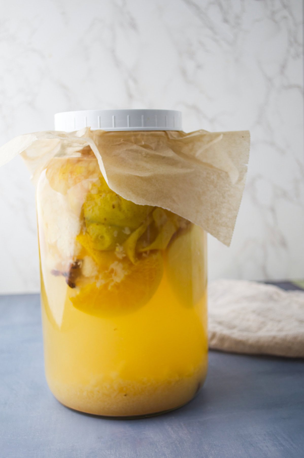 A large glass jar with a white lid and paper cover contains fermenting fruit pieces in yellow liquid, sitting on a blue surface with a beige cloth in the background.