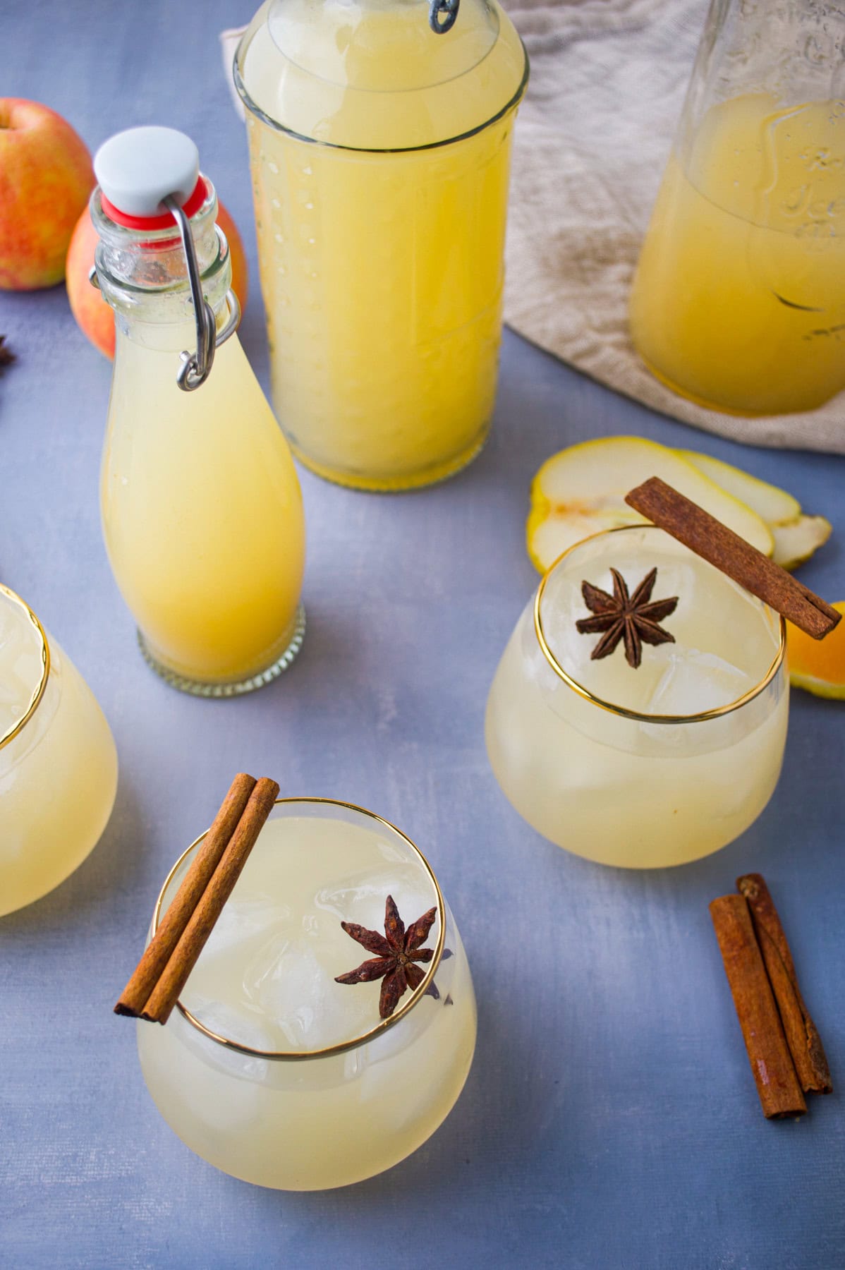 Three glasses of light yellow drink with ice, garnished with cinnamon sticks and star anise, are on a table with bottles of the same drink, fresh apple slices, and more cinnamon sticks nearby.