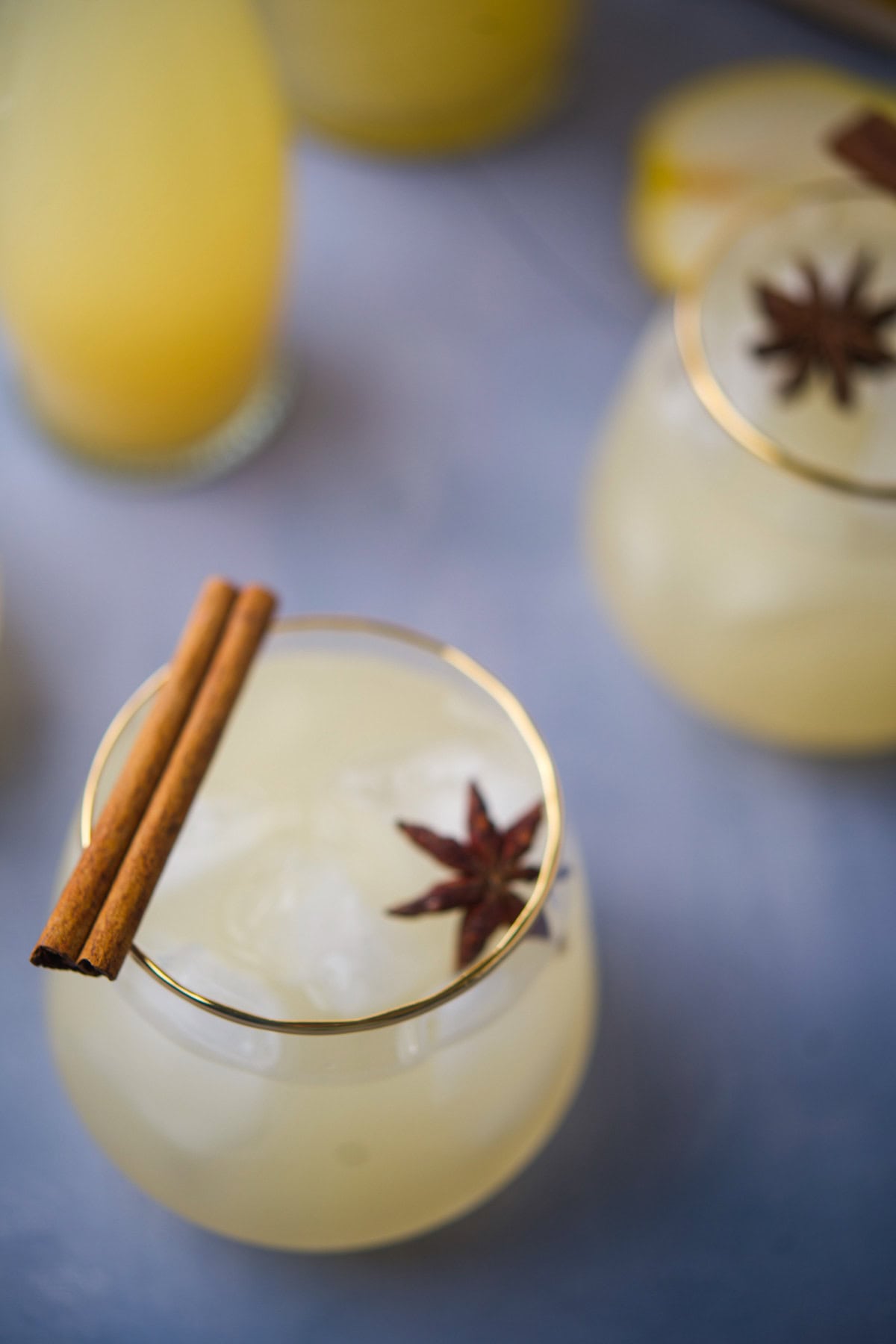 A glass of iced yellowish drink garnished with a cinnamon stick and star anise, with another similar drink and a glass of juice in the background. The glasses have gold rims and the scene is softly lit.