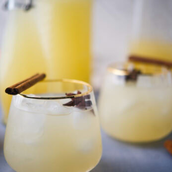 Two clear glasses filled with a DIY Fermented Apple Pear Soda (Apple Pear Kvass), drink, ice cubes, and garnished with cinnamon sticks. There are large glass bottles of the same drink in the background on a light surface.