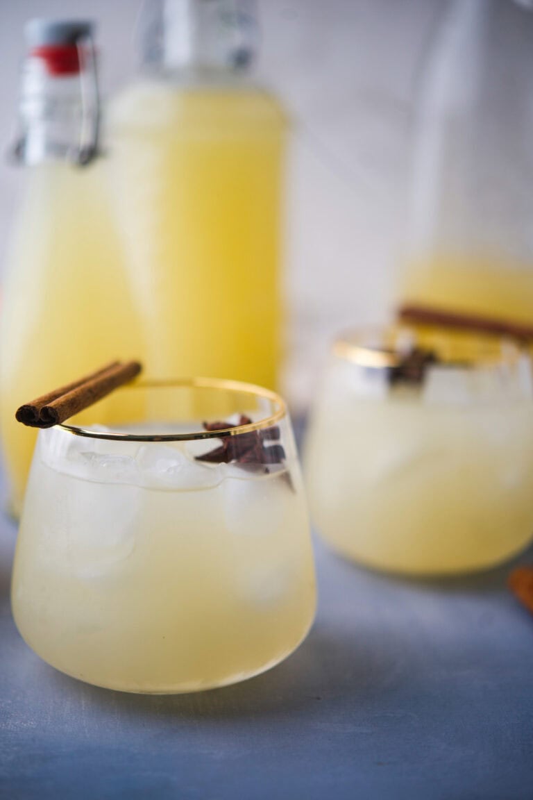 Two clear glasses filled with a DIY Fermented Apple Pear Soda (Apple Pear Kvass), drink, ice cubes, and garnished with cinnamon sticks. There are large glass bottles of the same drink in the background on a light surface.