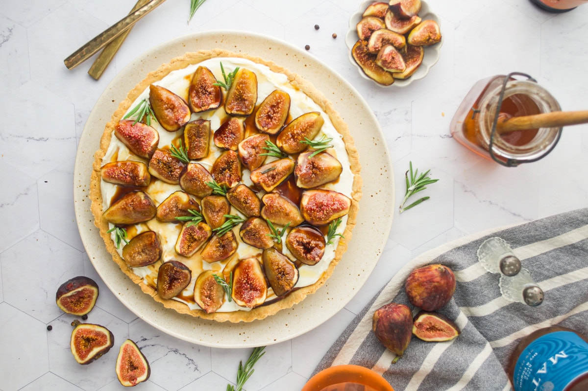 A fig tart topped with fresh fig slices and herbs sits on a plate, surrounded by whole and cut figs, a jar of honey, gold cutlery, and a striped cloth on a white marble surface.