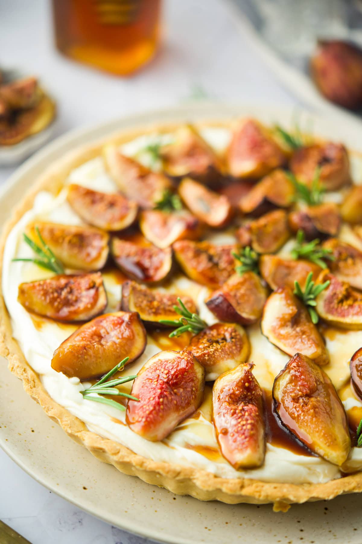 A tart topped with fresh figs, cream, and sprigs of rosemary sits on a plate. The figs are sliced and arranged over a creamy filling in a golden tart crust. A honey jar and more figs are blurred in the background.