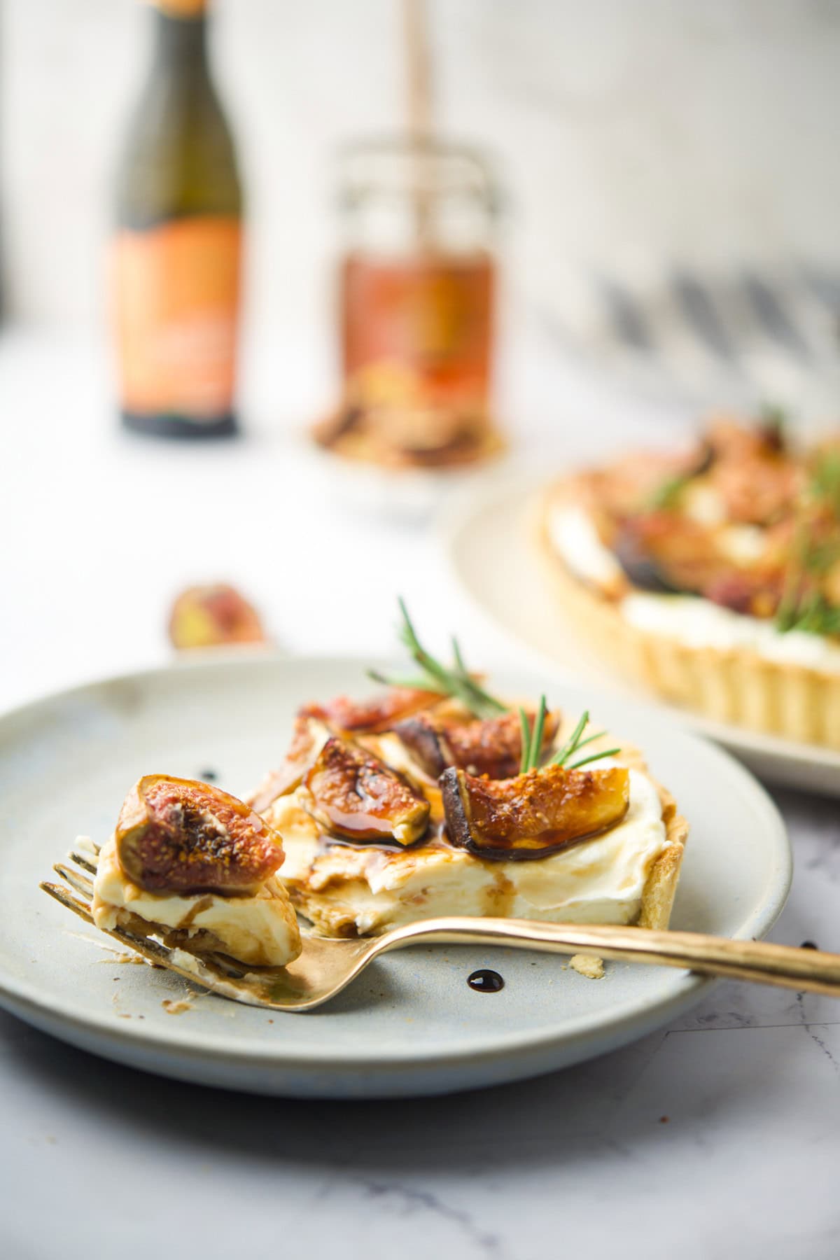 A slice of tart topped with fresh figs, cream, and rosemary sits on a gray plate with a fork. A bottle and a jar are blurred in the background, along with another tart on a plate.