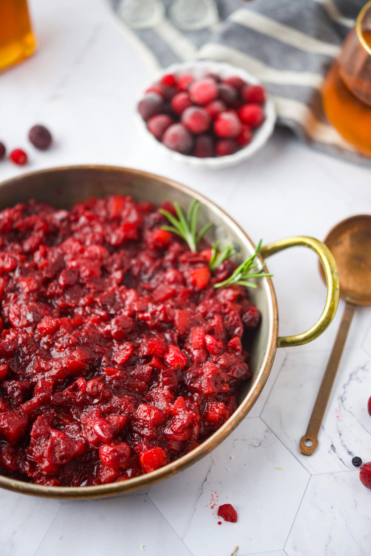 A bowl of chunky cranberry apple sauce garnished with a sprig of rosemary is shown on a white surface, with a spoon nearby and a small bowl of fresh cranberries in the background.