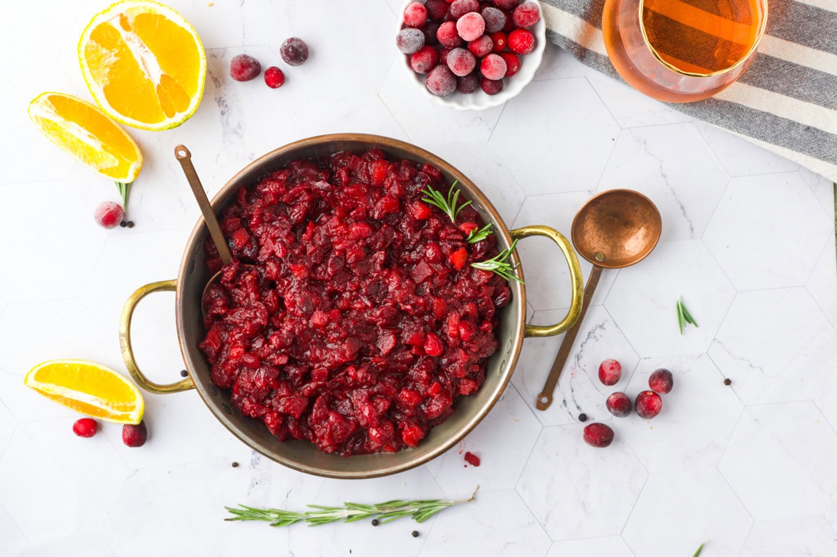 A bowl of cranberry sauce with a serving spoon, surrounded by fresh cranberries, rosemary sprigs, orange slices, and a glass of orange liquid on a white marble surface.