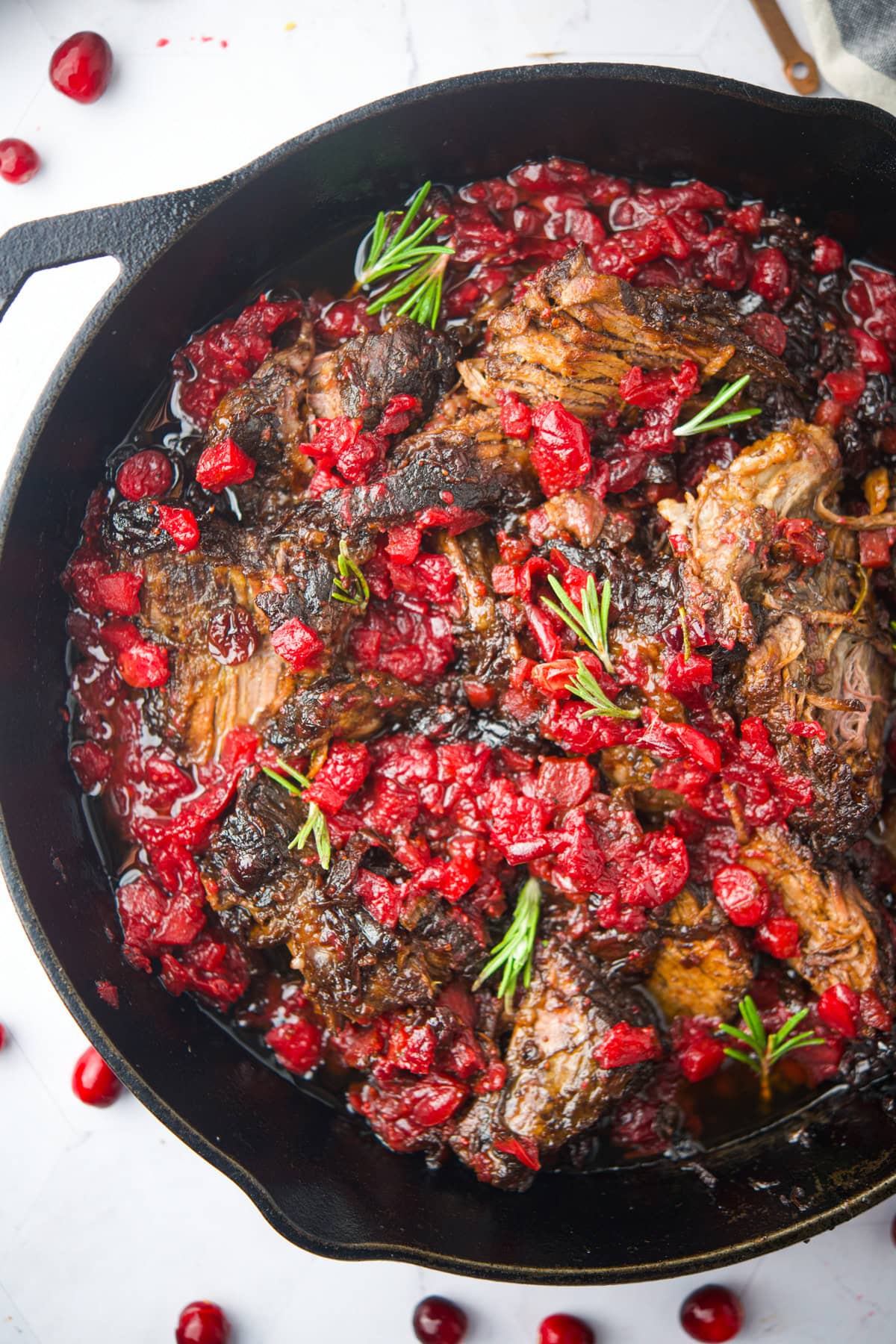 Melting pot of braised beef with herbs, red wine, and fresh cranberries in a cast iron skillet.