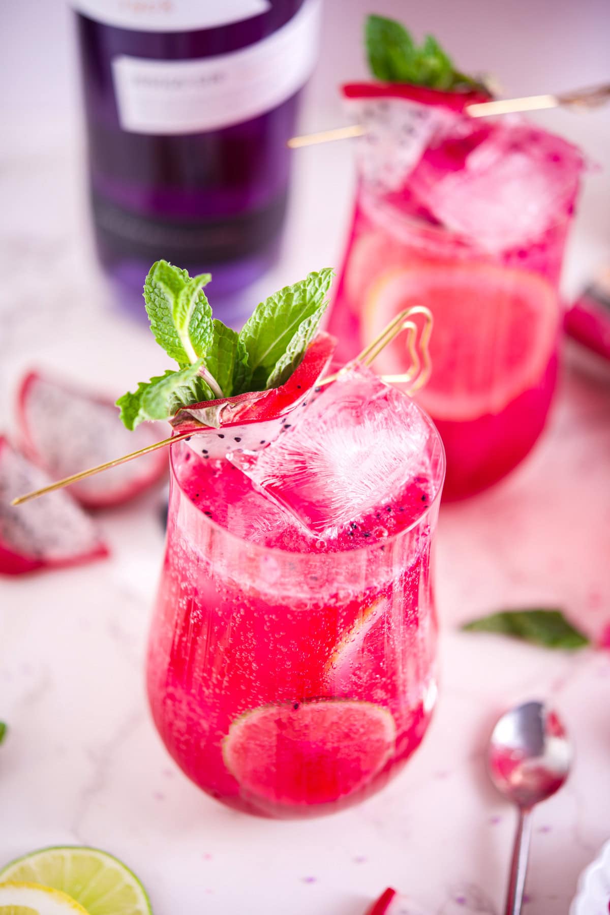 A vibrant pink dragon fruit cocktail in a clear curved glass garnished with a slice of lemon, mint leaves, and a skewer of dragon fruit. The drink is fizzy and refreshing, with visible bubbles and dragon fruit seeds. A purple bottle is blurred in the background, and lemon and dragon fruit slices are scattered on the marble surface.