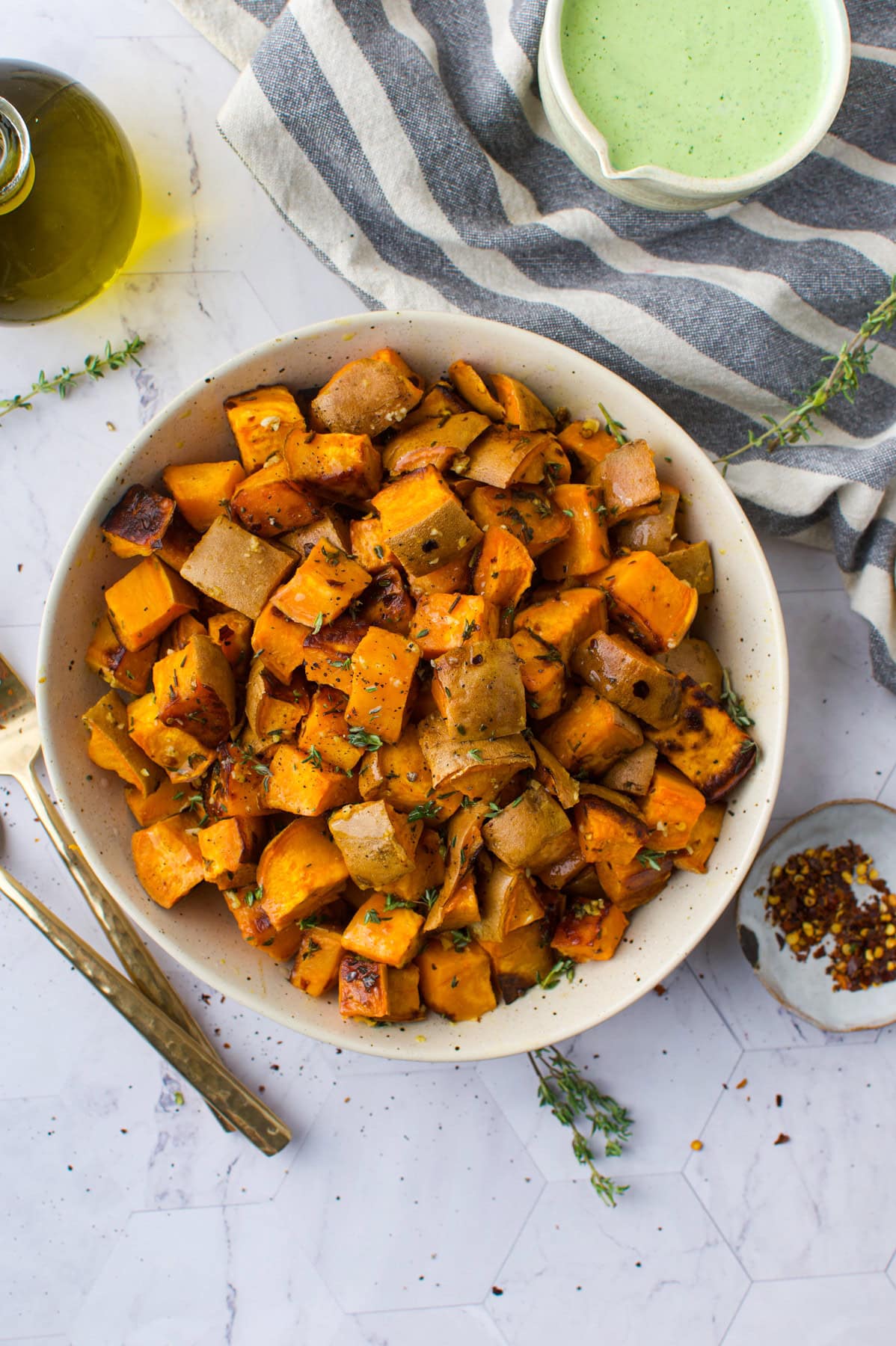 A bowl of roasted sweet potato and potato cubes garnished with herbs sits on a white surface next to gold utensils, a striped cloth, a small bowl of green sauce, olive oil, and red pepper flakes.