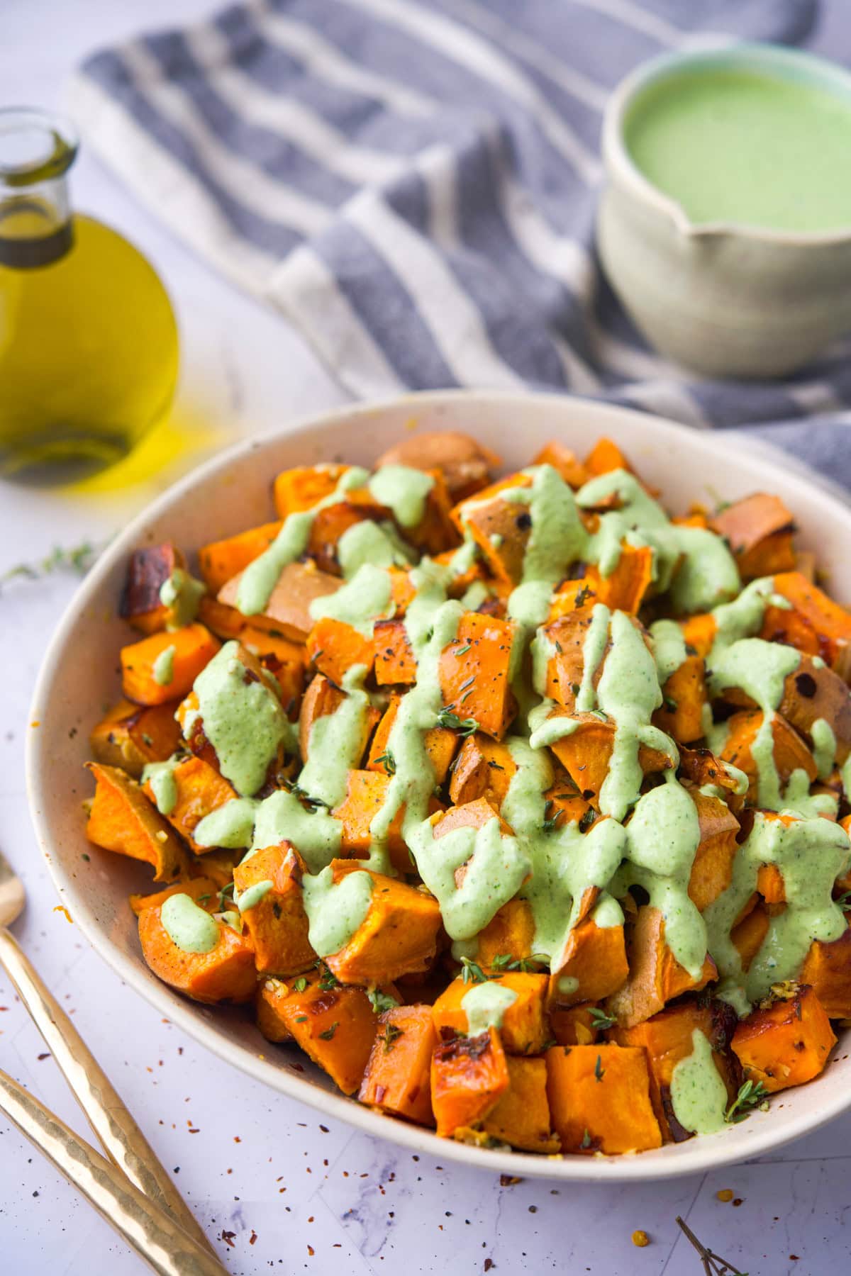 A bowl of roasted sweet potato cubes topped with a green herb sauce, set on a table with a striped cloth, a small bottle of olive oil, and a bowl of extra sauce in the background.