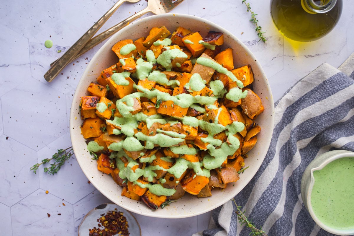 A bowl of roasted sweet potatoes topped with a creamy green sauce, surrounded by utensils, a striped towel, and a bottle of olive oil on a marble surface.